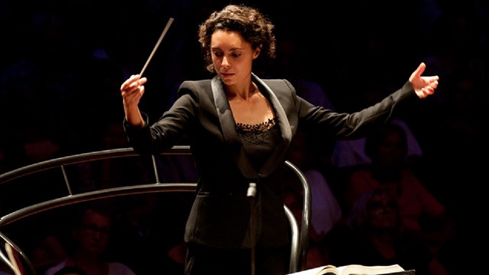 A female alumna, with dark brown hair, wearing a smart black suit, conducting from a conductors podium.